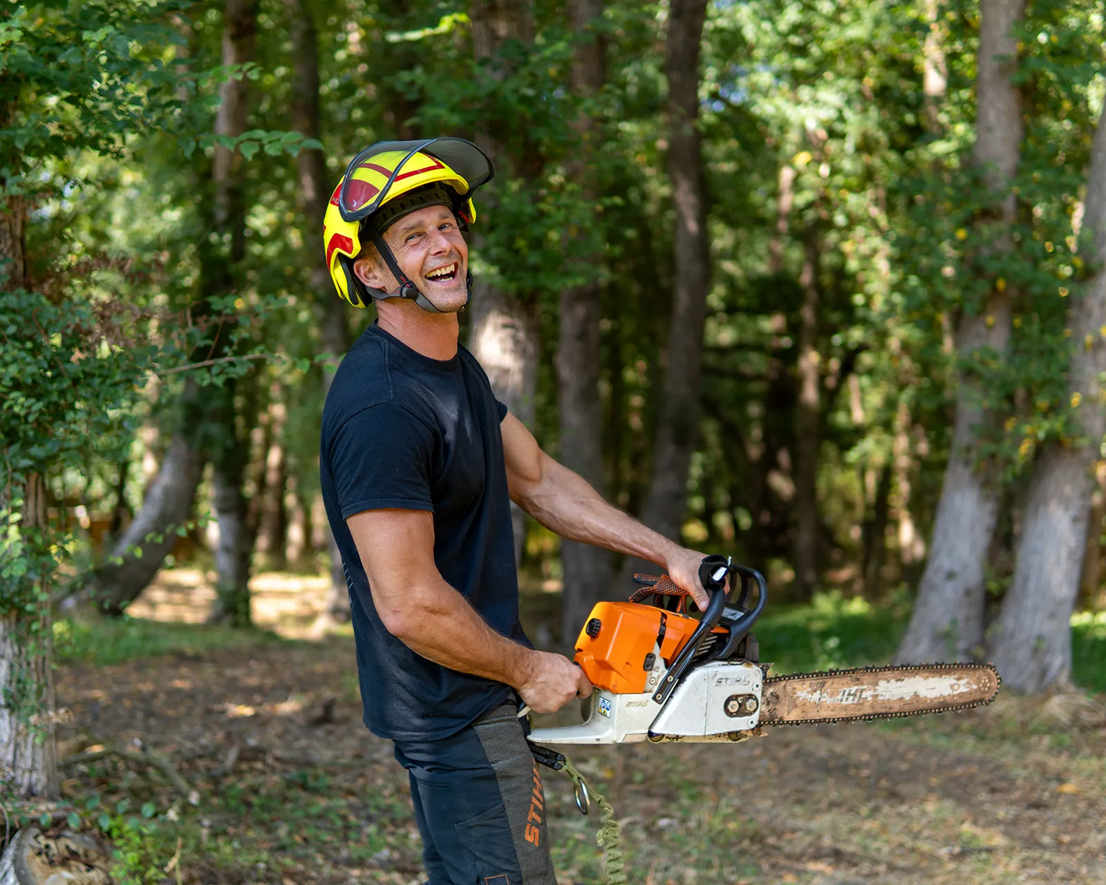 Man in safety gear holding a chainsaw, smiling in a forest setting with trees in the background.